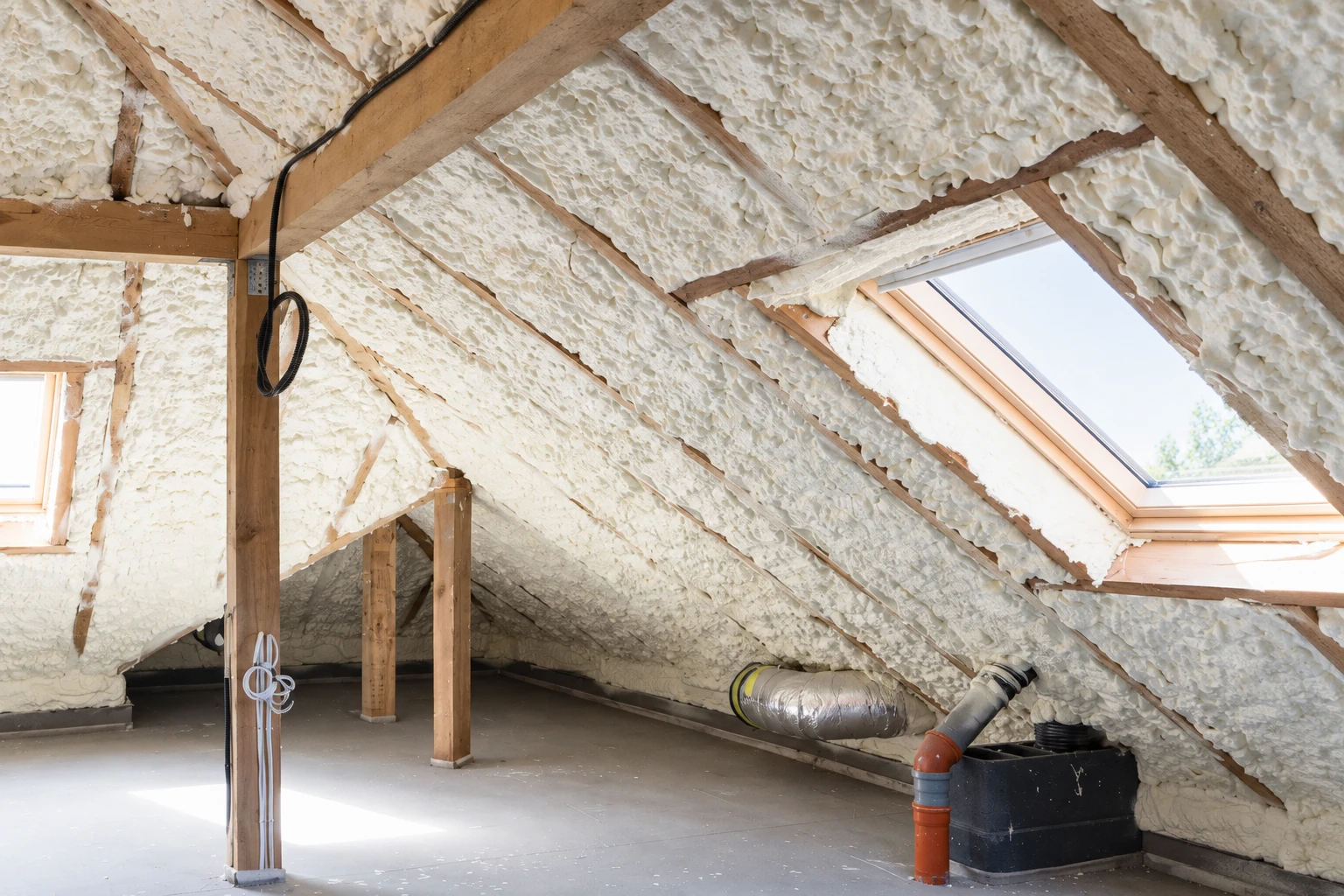 Attic rafters and roof slopes coated with white spray foam, with a skylight and ductwork visible.