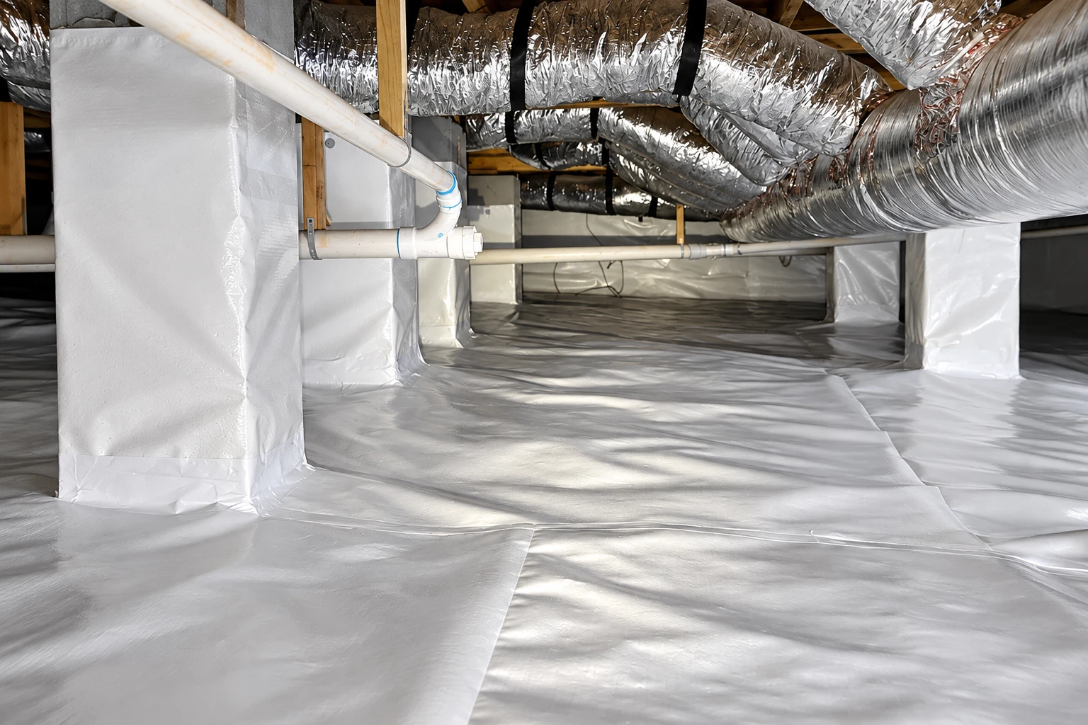 A crawl space with a white vapor barrier covering the floor and supports beneath overhead silver HVAC ducts and pipes.