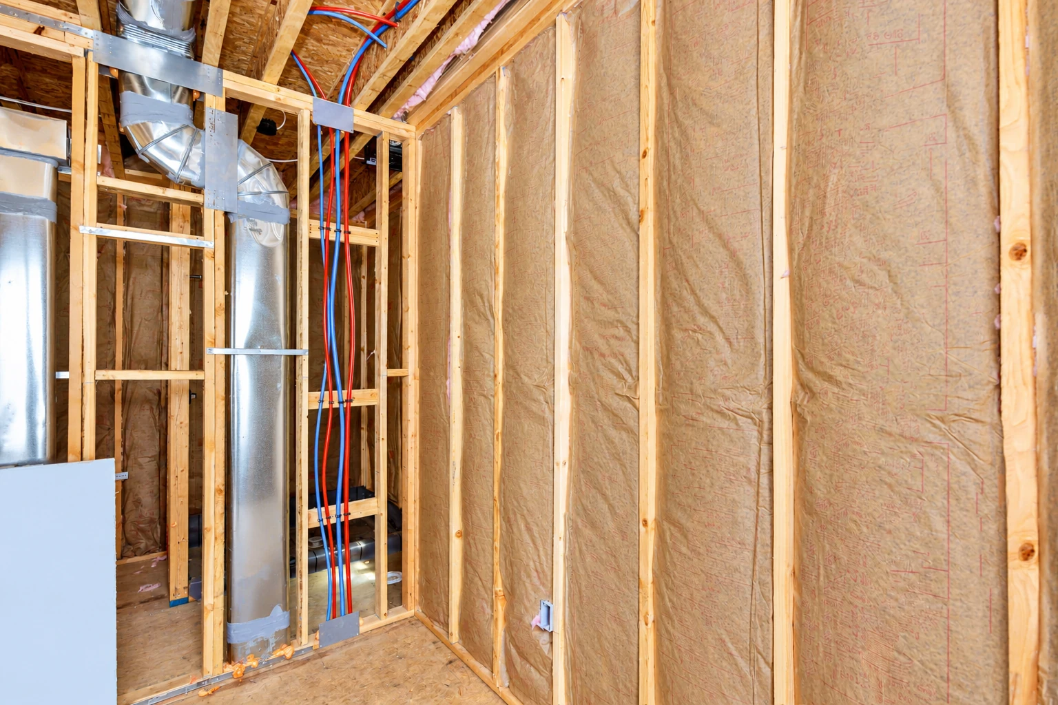 Fiberglass batts fill the wall cavities of an unfinished home beside exposed ducts and red and blue pipes.