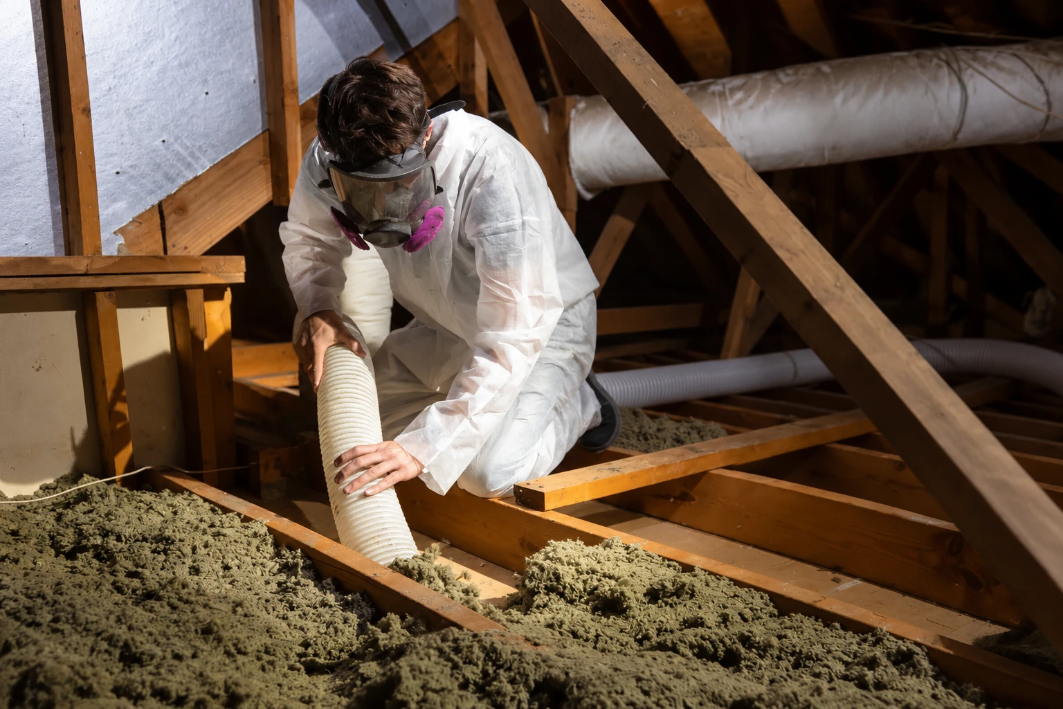 A worker in protective coveralls and a respirator uses a flexible hose to remove loose attic insulation.