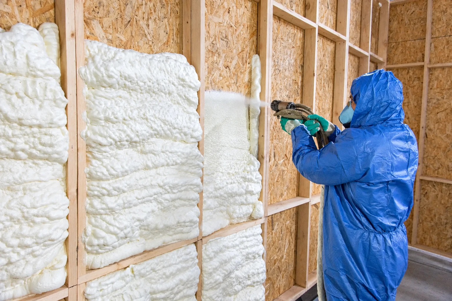 A worker in blue protective gear sprays white foam insulation into the exposed wooden wall cavities of a building.