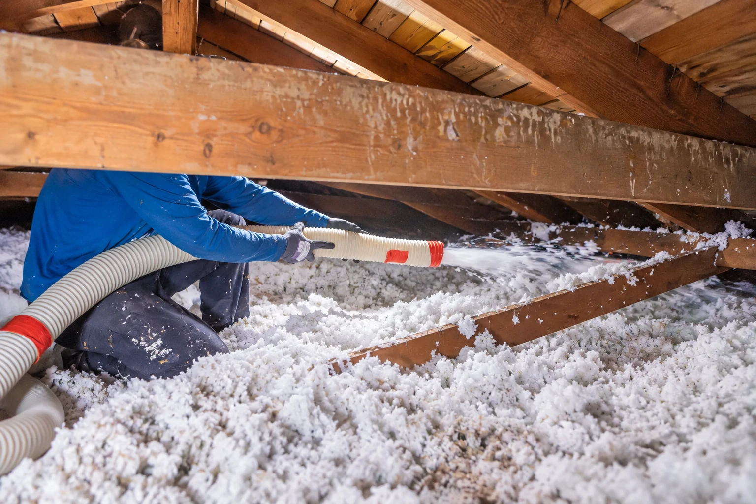 A worker directs a flexible hose to blow white insulation across the floor of a timber attic.