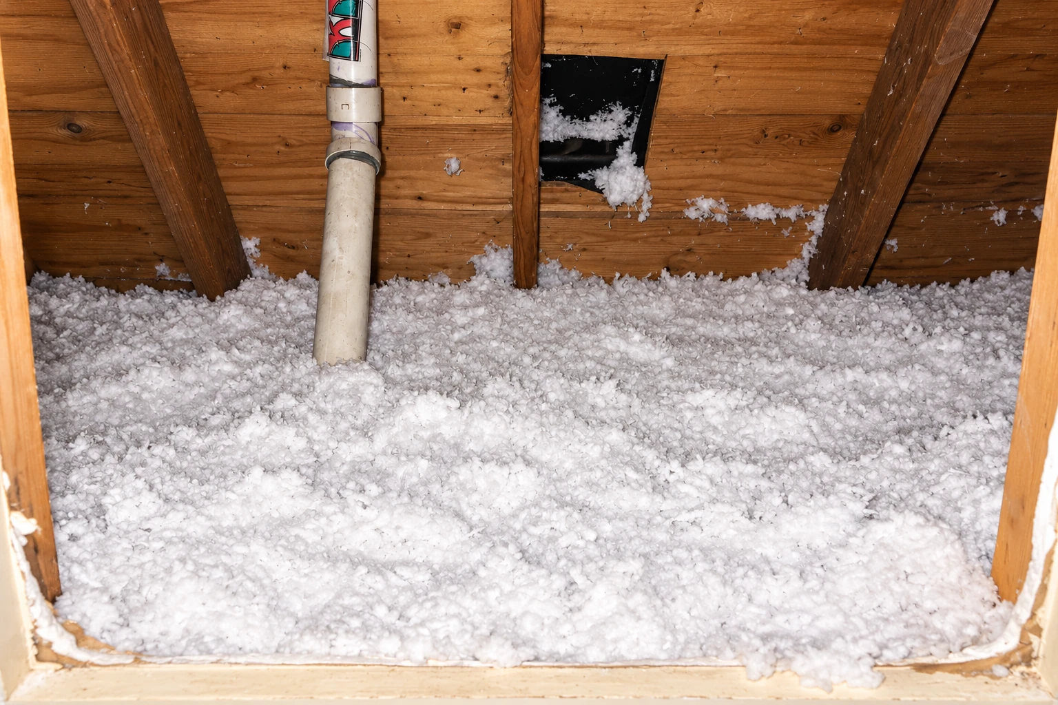 White blown-in insulation fills an attic behind a small access opening, with a pipe running through it.