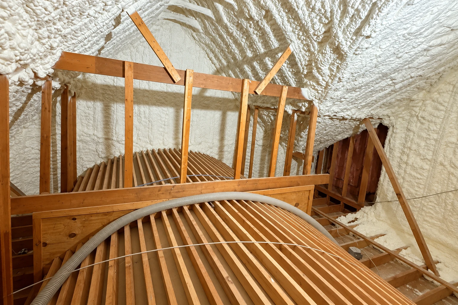 Spray foam insulation coats the sloped roof and walls of an attic around exposed wooden framing and a loft platform.