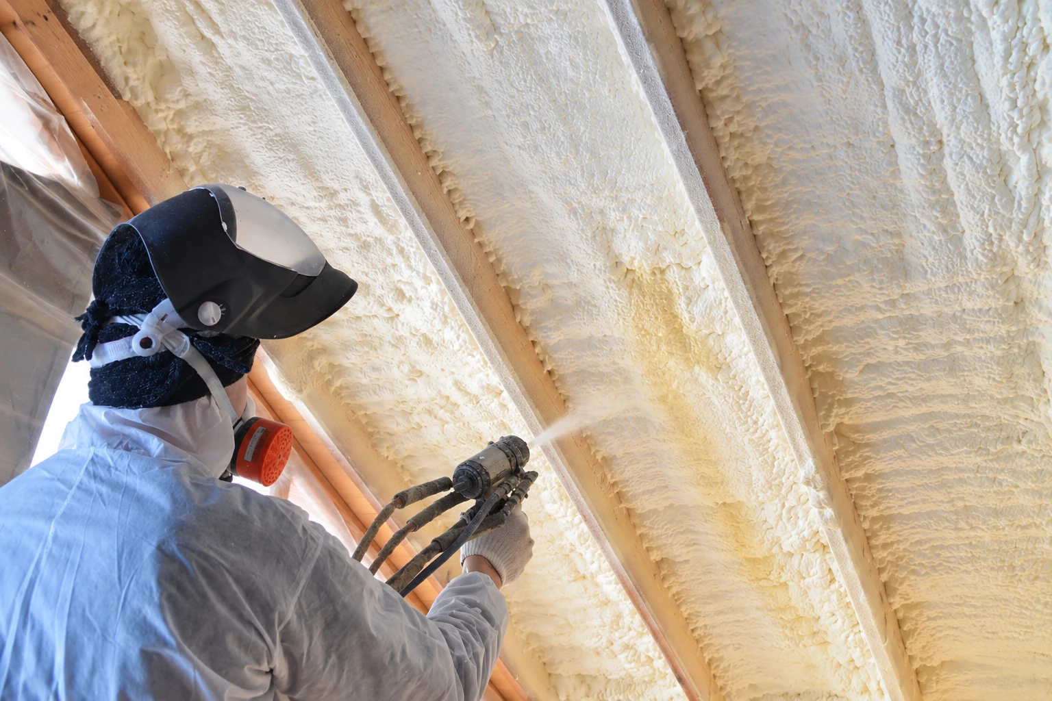 Worker in protective gear sprays foam insulation into the ceiling bays between exposed wooden rafters.