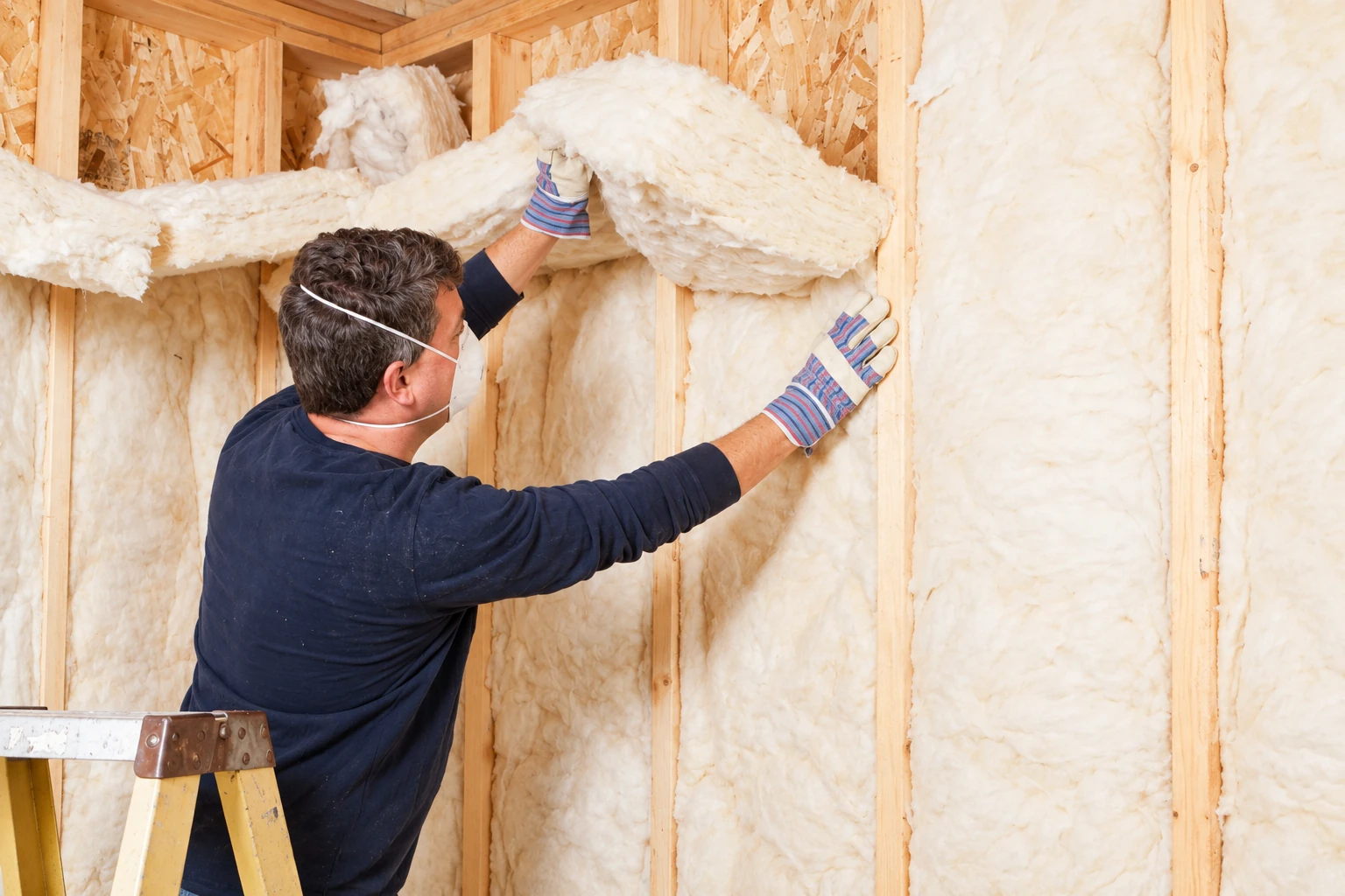 Worker in a mask and gloves fitting fluffy fiberglass insulation between wooden wall studs.
